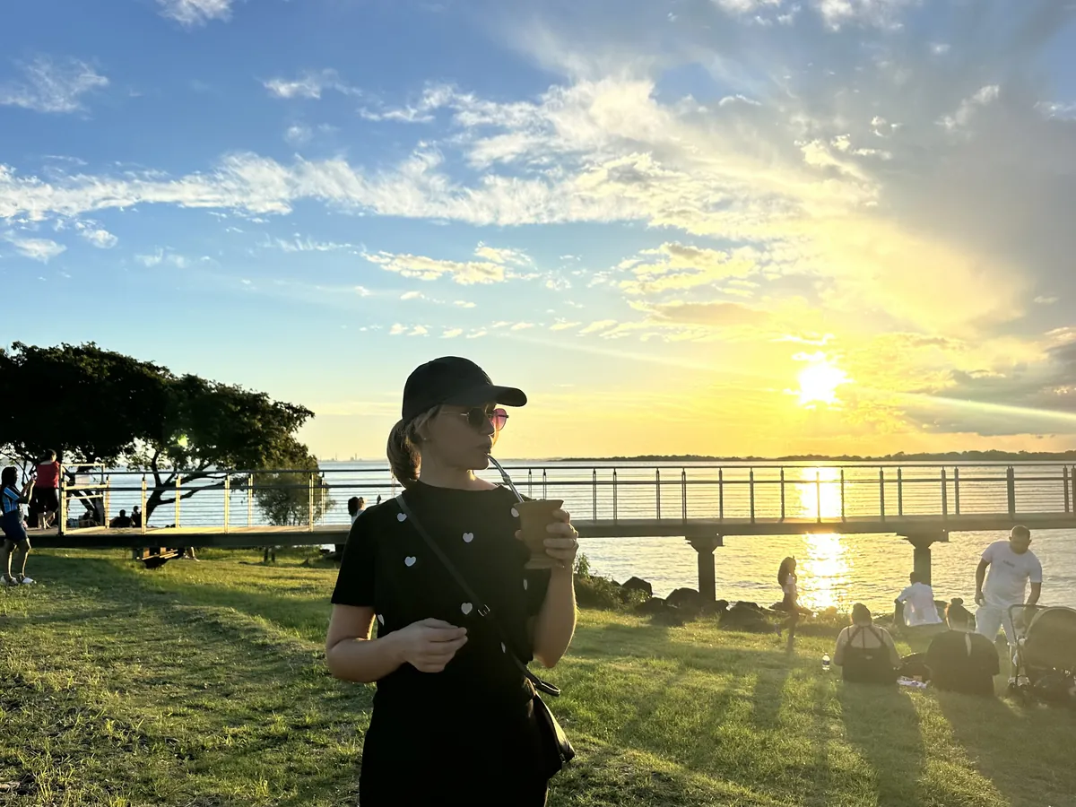 Ana drinking chimarrão from a gourd at sunset on a grassy waterfront, a pier and the setting sun behind her