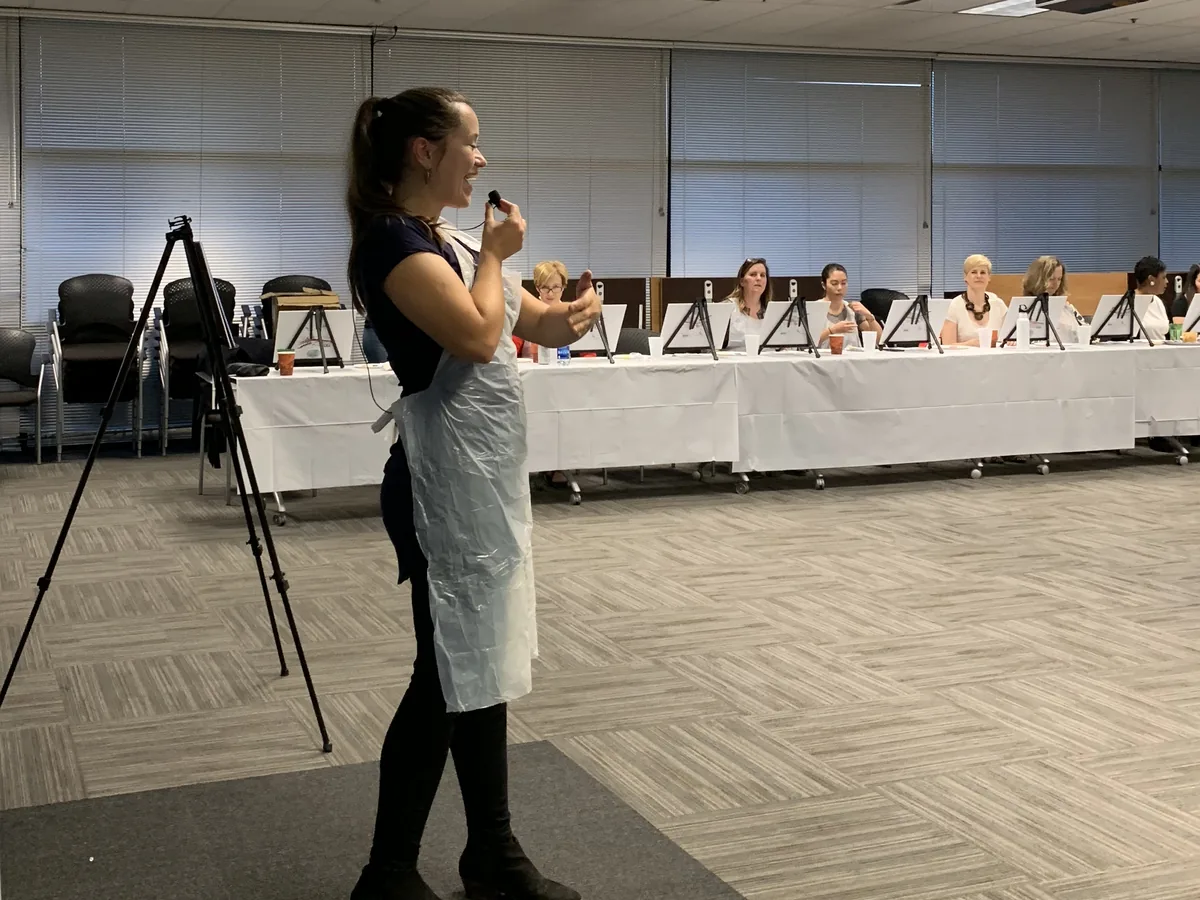 Ana teaching with a microphone at the front of a company conference room, the team at their easels behind her