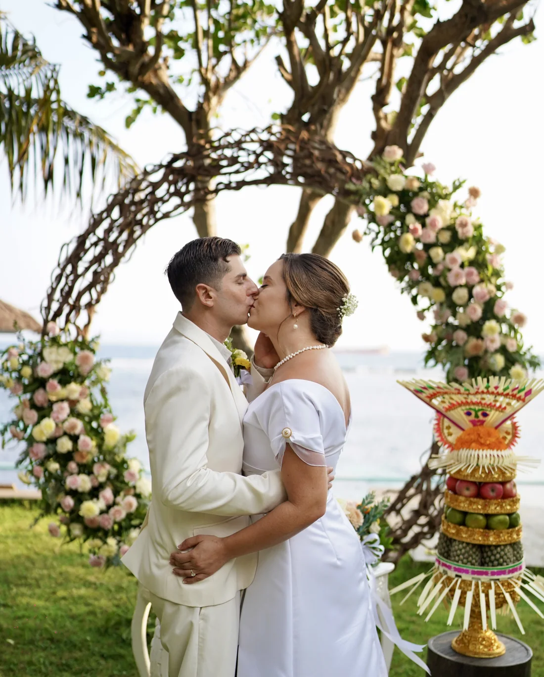 Ana and her husband kissing at their wedding in Bali, beside a tall Balinese fruit offering and an arch of roses by the sea